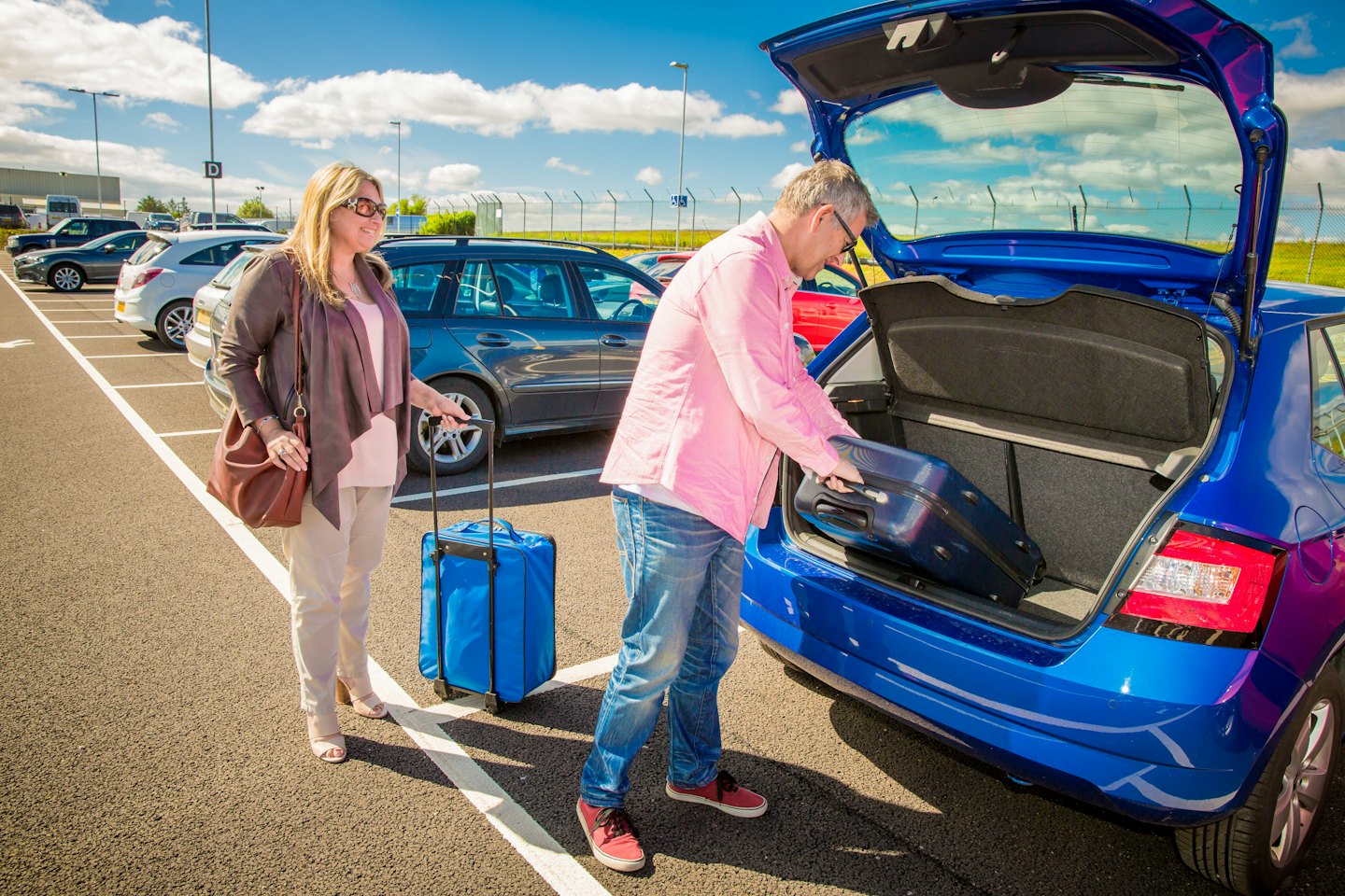 Man loads suitcase into blue car trunk with woman and luggage.